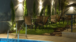 Una zona junto a la piscina con piscina, sillas y mesas, rodeada de vegetación y muros con luces.
