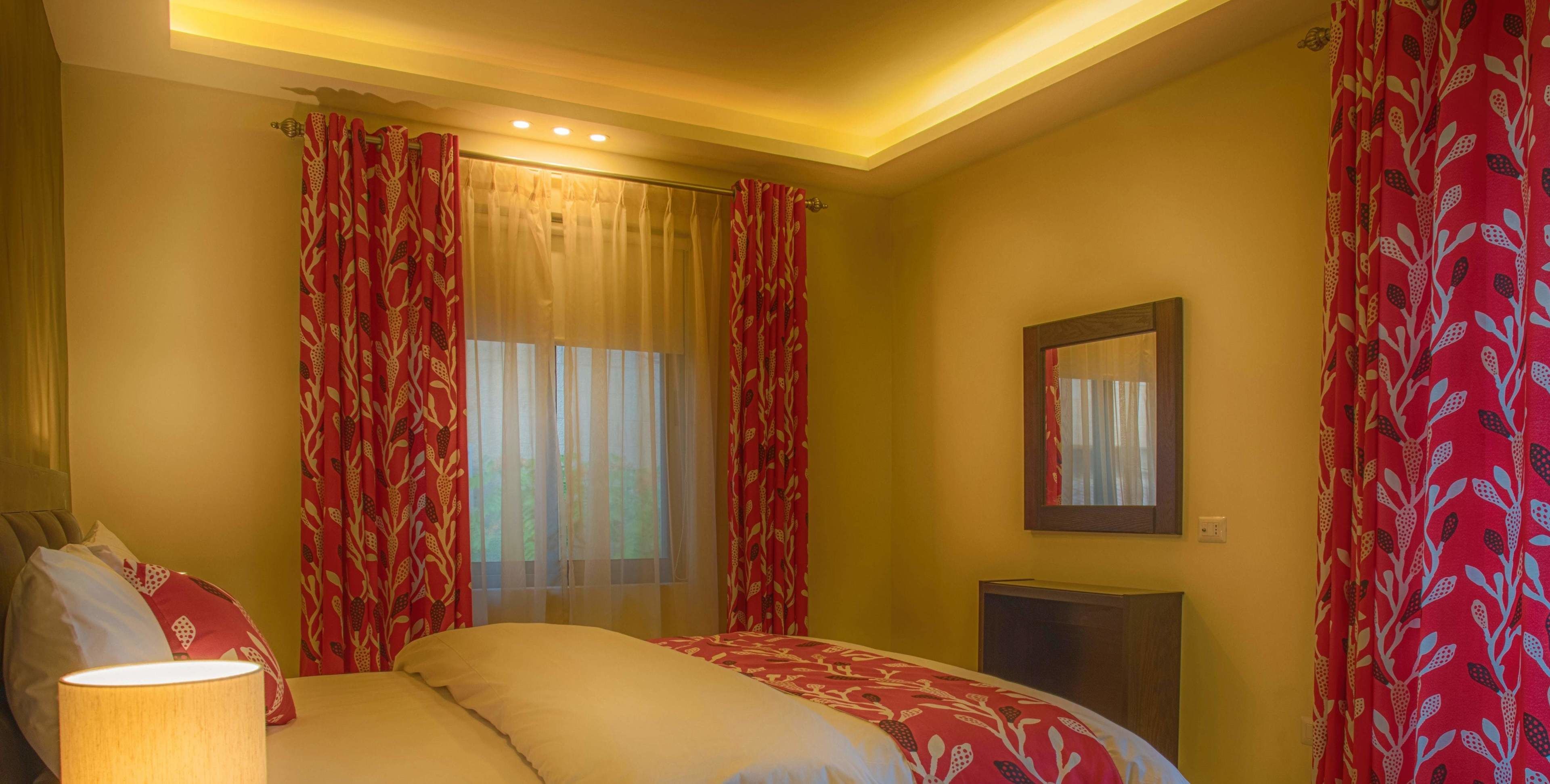 A bedroom with a bed, a bedside lamp, a mirror, and red patterned curtains.