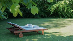 A wooden lounge chair with a mattress and rolled towels on a grassy lawn, surrounded by dense foliage. - B&B Scheldekant (Oudenaarde)