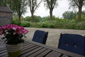 Et træbord med to stole, en potteplante og en grussti, der fører til et træhegn og træer.