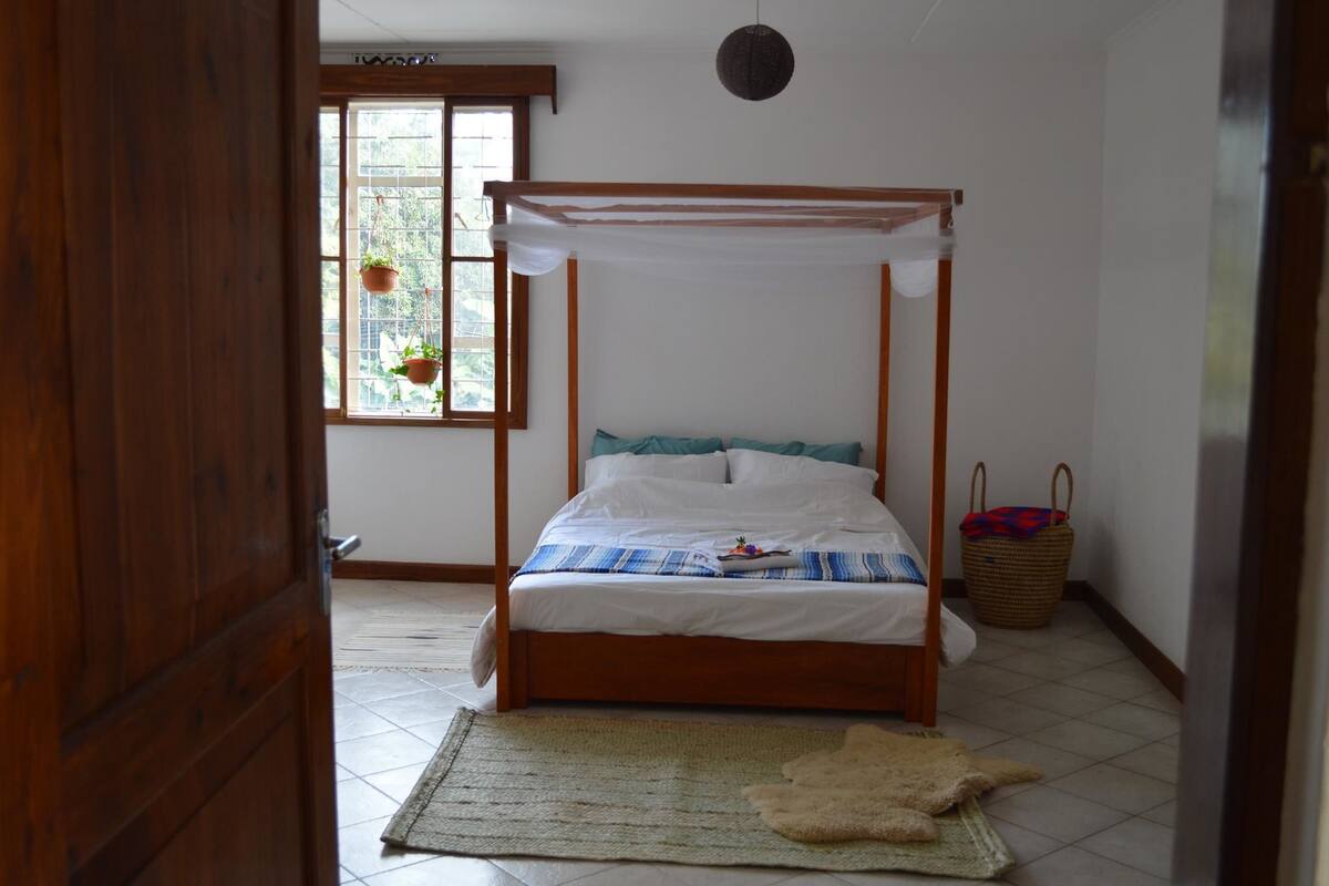 A bedroom with a wooden four-poster bed, a window with potted plants, a woven rug, and a woven basket.