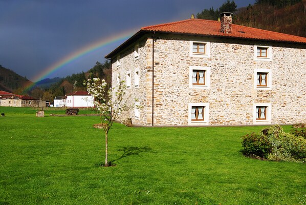 A stone building with a red roof, surrounded by a green lawn and a rainbow in the background.
