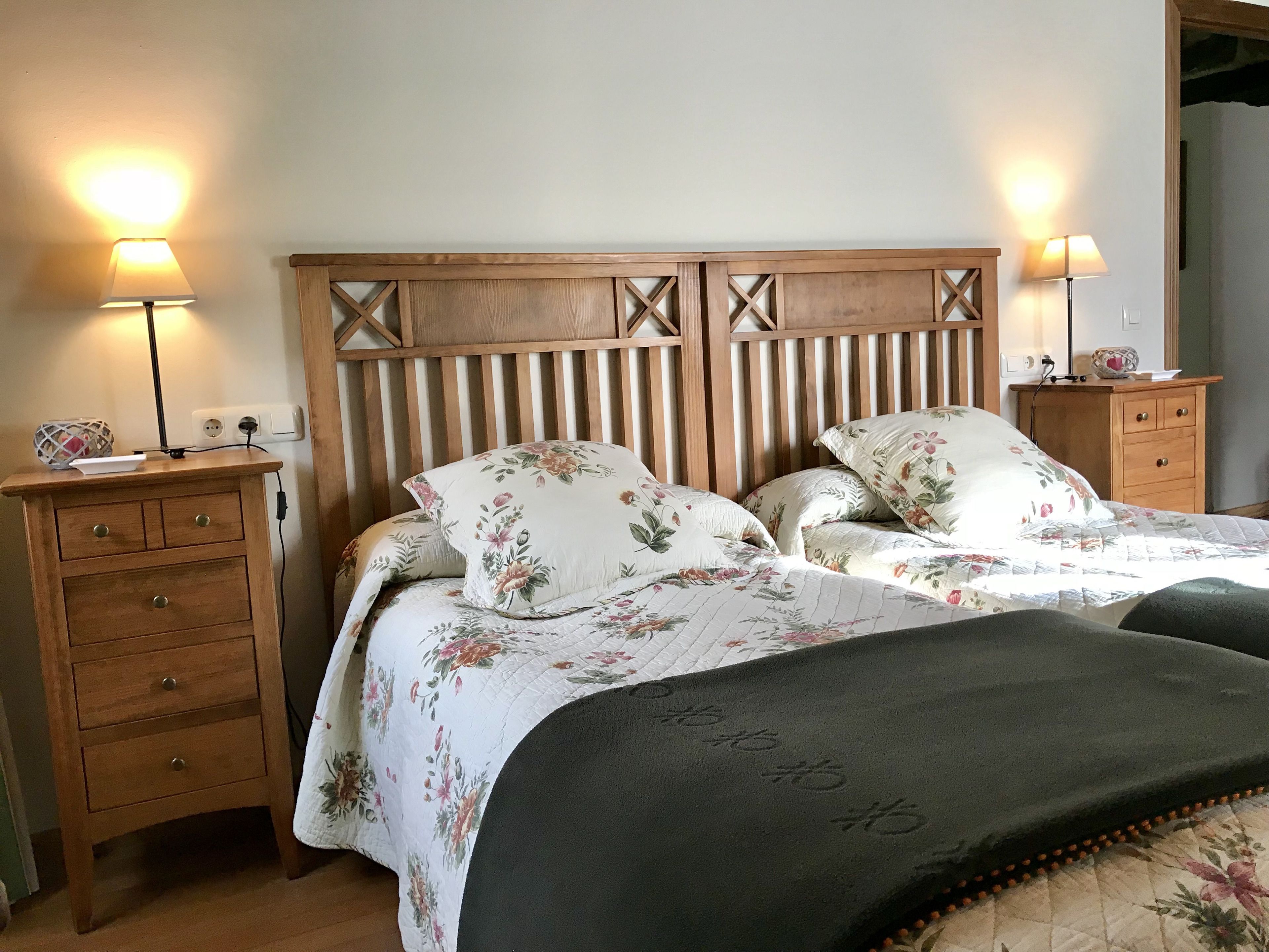 Un dormitorio con cabecera de madera, ropa de cama floral, dos mesitas de noche con lámparas y una pequeña mesita de noche con lámpara.