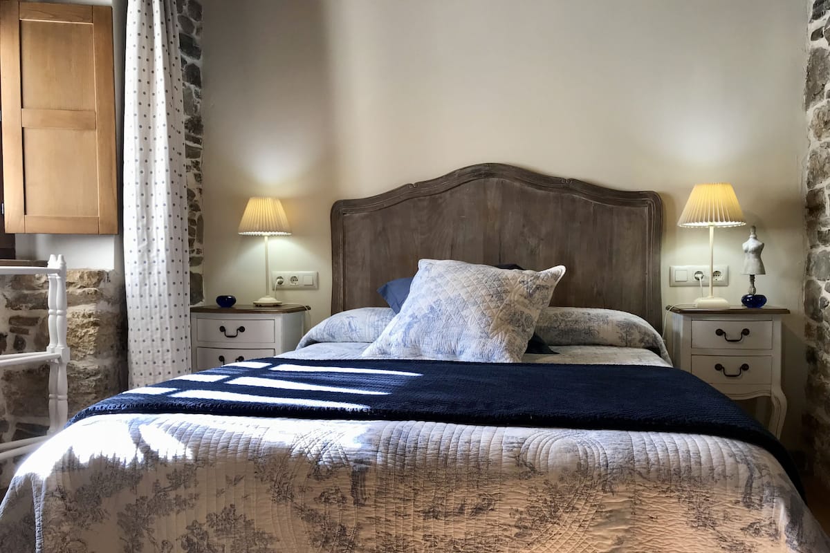 Un dormitorio con cabecera de madera, dos mesitas de noche con lámparas, una cama con ropa de cama estampada y una pared de piedra.
