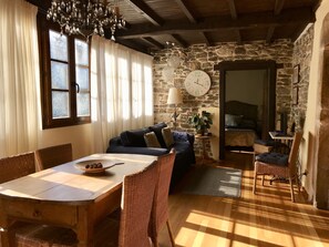 A cozy room with a stone wall, a wooden ceiling, a dining table with chairs, a sofa, and a large clock on the wall. - Ca Lulón (Tineo)