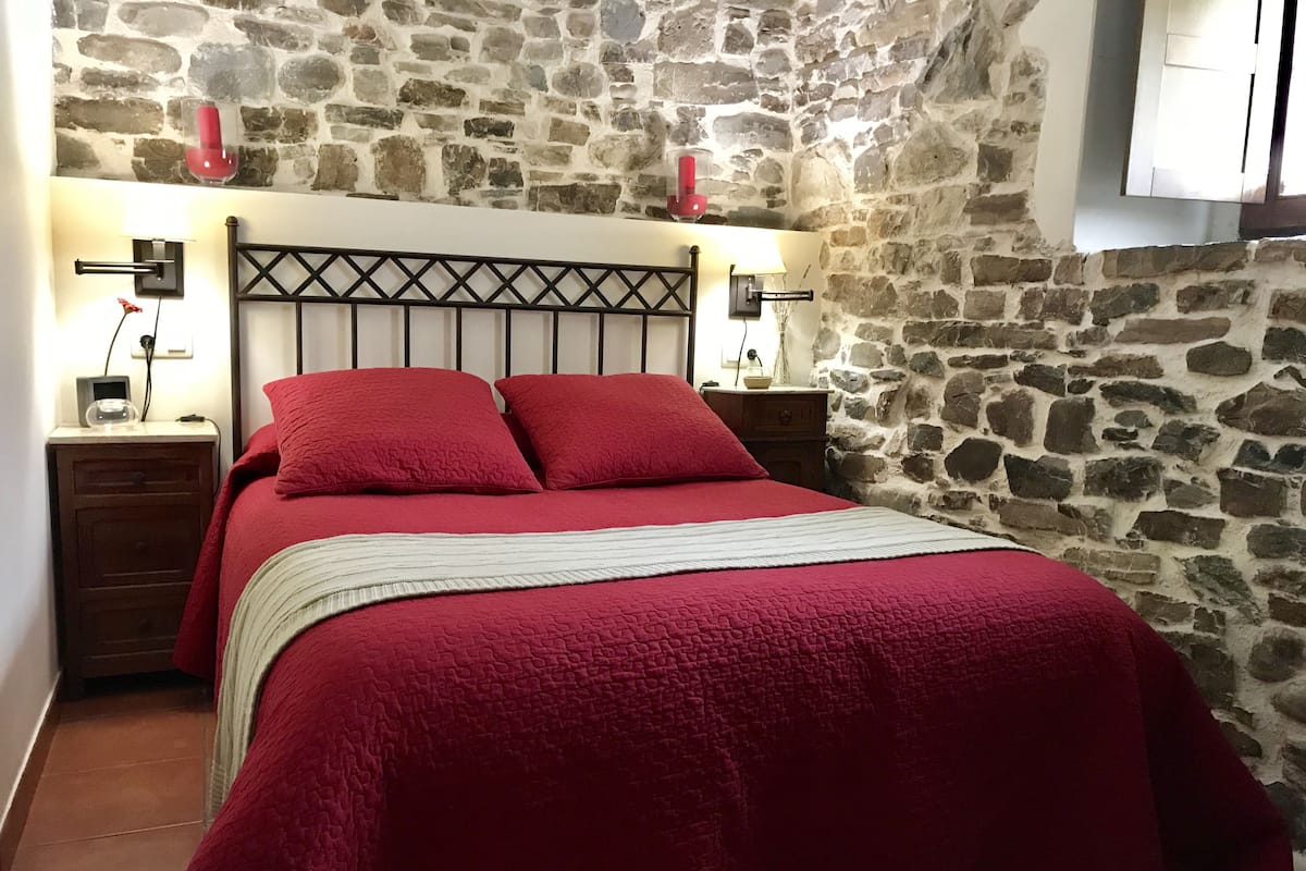 Un dormitorio con pared de piedra, una cama con ropa de cama roja, dos mesitas de noche con lámparas y una mesita con un reloj.