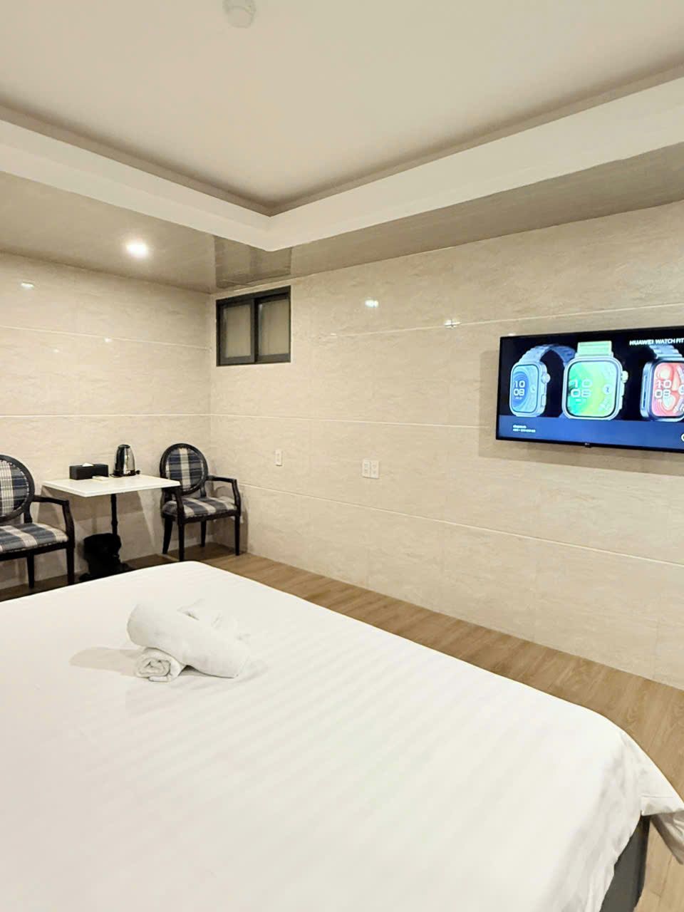 Una camera d'albergo con un letto, un tavolino con sedie e una TV a muro che mostra una pubblicità di orologi.