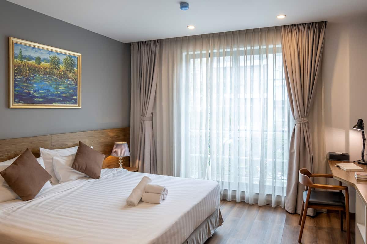 Una cama bien hecha con almohadas, un escritorio con una silla y un ventanal con cortinas.
