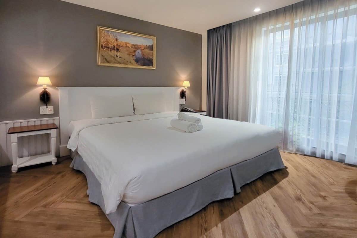 Una cama grande con ropa blanca, dos mesitas de noche con lámparas, un cuadro en la pared y una ventana con cortinas.