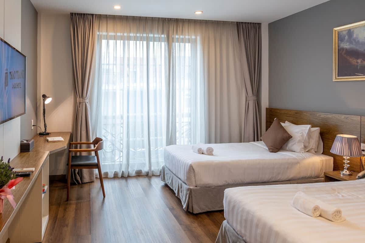 Habitación de hotel con dos camas, un escritorio, una silla y un ventanal con cortinas.