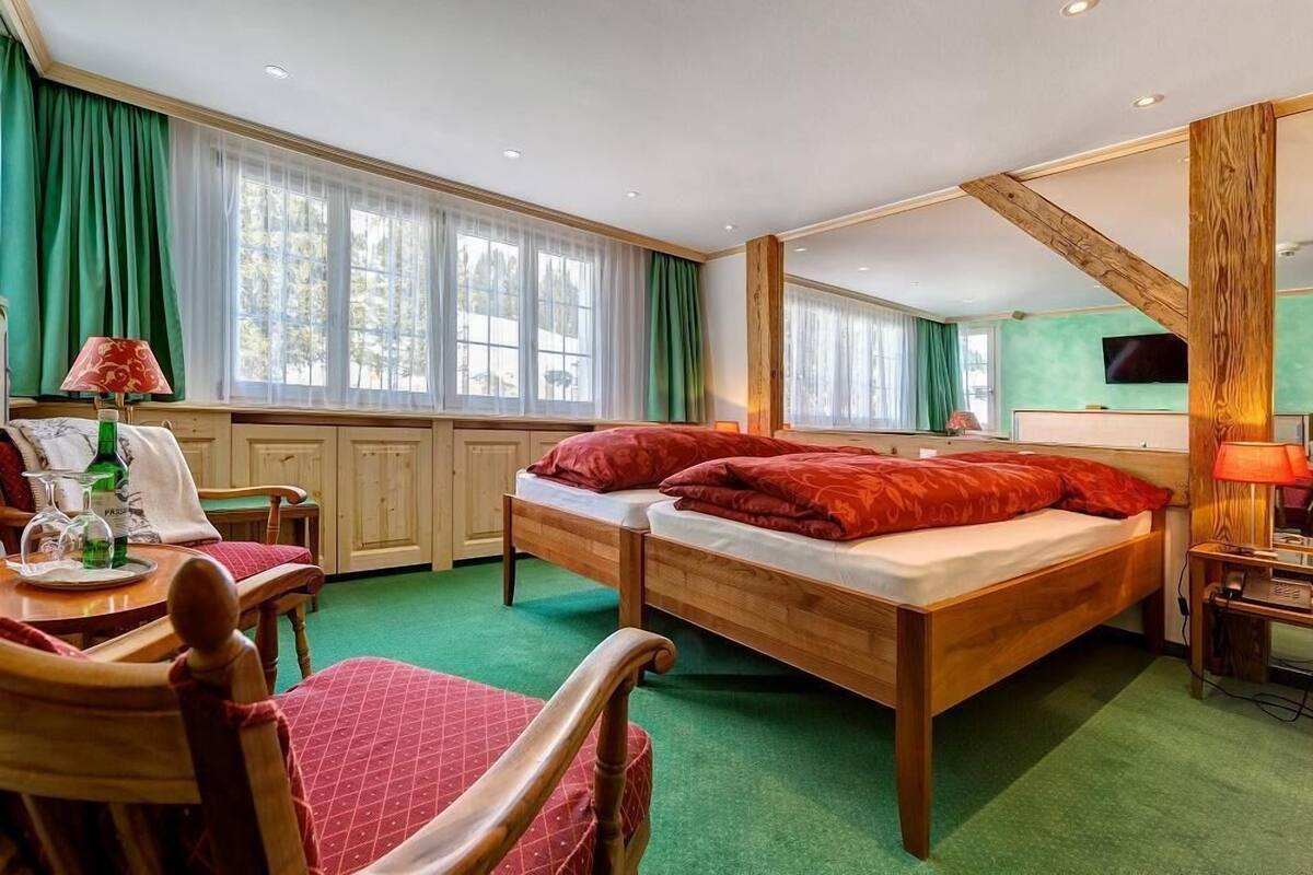 A hotel room with two wooden beds, a green carpet, a wooden chair, a table with a lamp, and a TV on the wall.