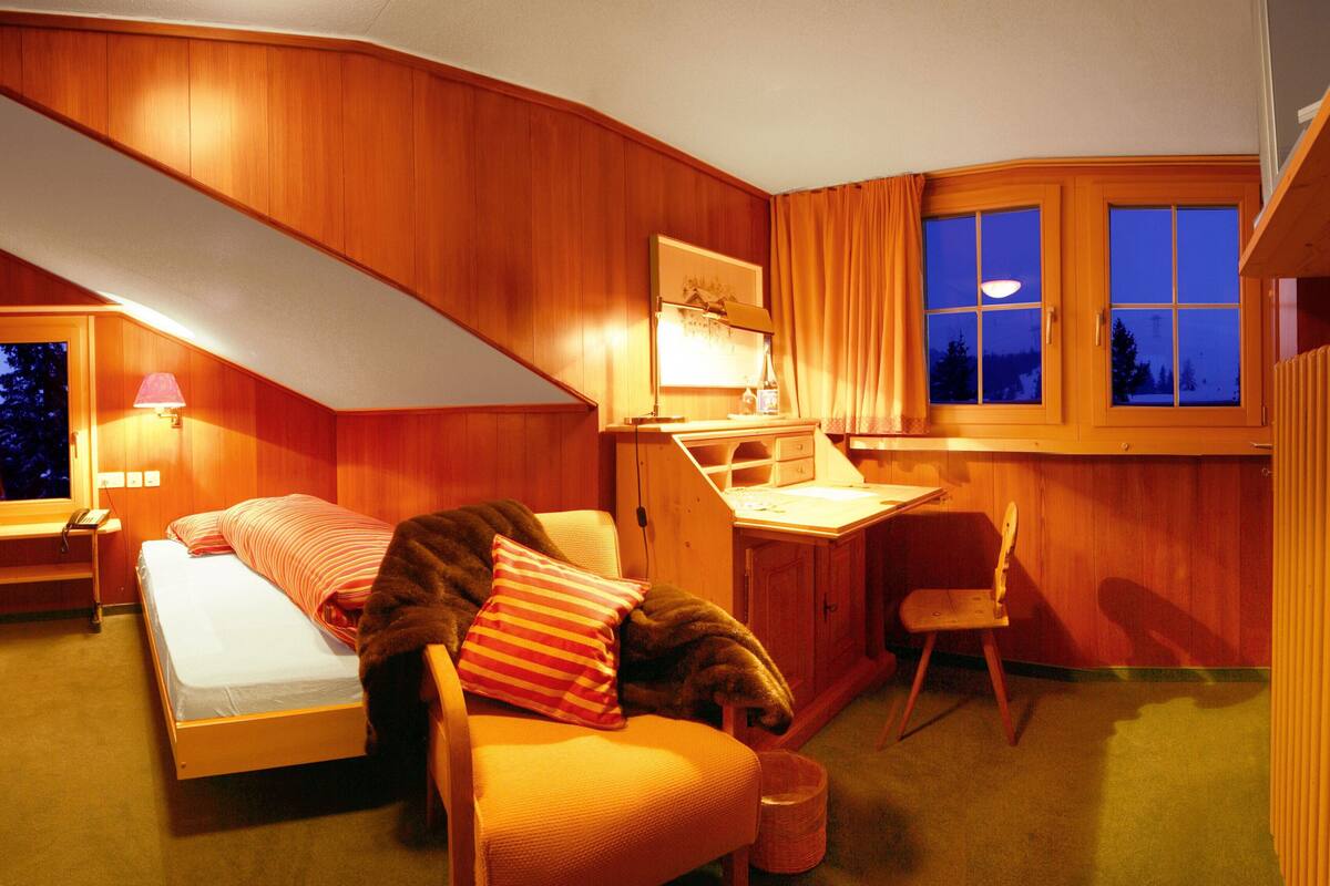 A cozy attic bedroom with a bed, desk, and chair, wooden walls, and a window with curtains.