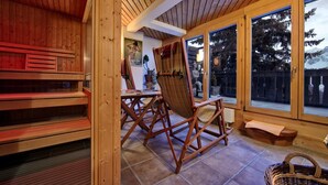 A wooden interior with a sauna, a dining area with chairs, and large windows offering a view of trees and mountains. - Hotel Arlenwald (Arosa)