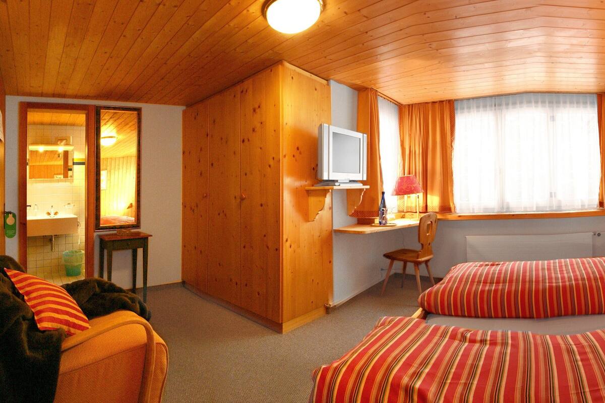 A cozy room with wooden walls, a bed with striped bedding, a desk with a chair, and a window with curtains.