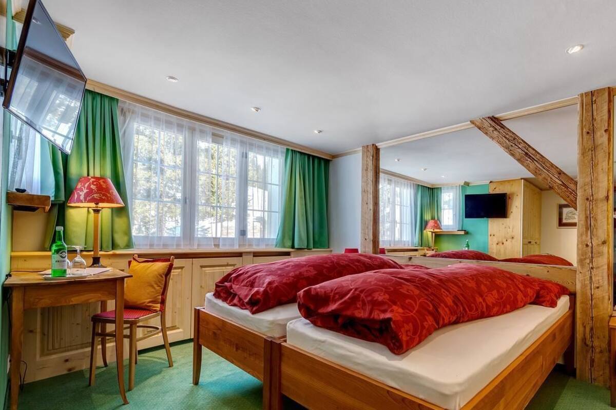 A room with two wooden beds, a desk with a chair, a television, and large windows with green curtains.
