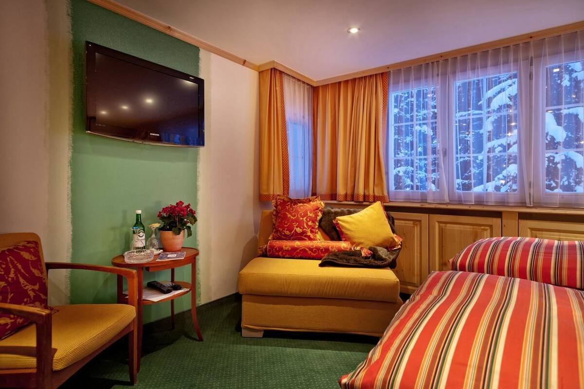 A hotel room with a sofa, a chair, a TV mounted on the wall, a window with curtains, and a view of snow-covered trees.
