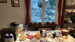 A breakfast table set with various food items and drinks, a coffee maker, and a window with a view of a garden. - Bed & Breakfast Obermumpf (Obermumpf)