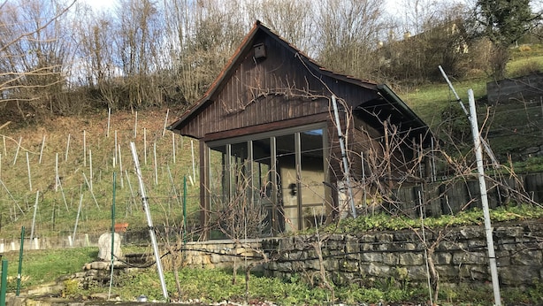 A rustic wooden building with a sloped roof, surrounded by a stone wall and a garden with grapevines. - Bed & Breakfast Obermumpf (Obermumpf)