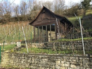 Un bâtiment rustique en bois, doté d’un toit en pente, entouré d’un mur de pierre et d  ’un jardin où poussent des vignes.