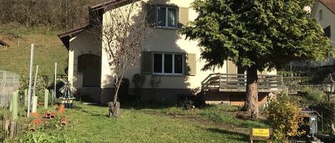Una casa de dos pisos con techo inclinado, una chimenea visible y un pequeño jardín con un árbol y césped.