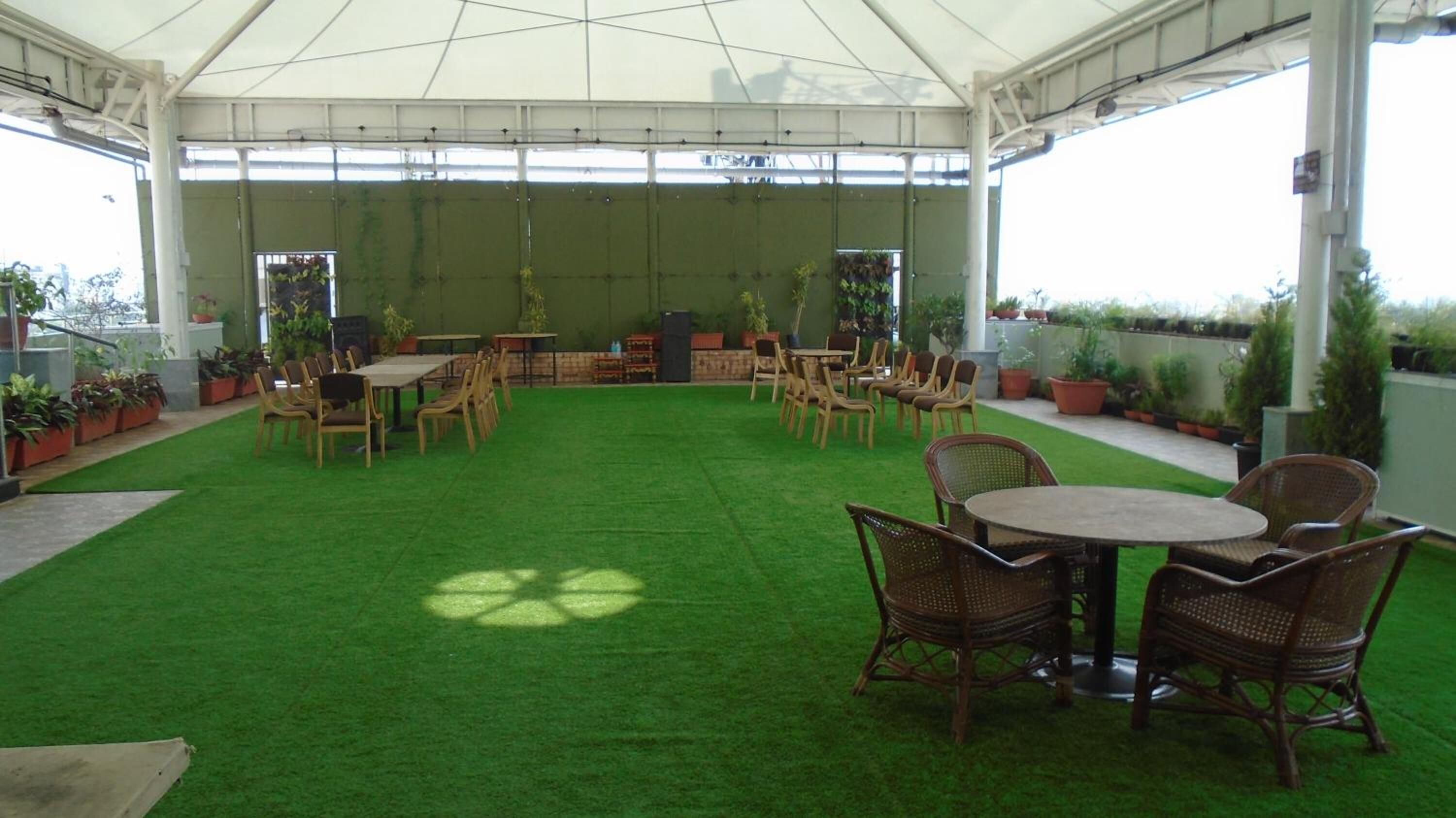 A covered outdoor patio with artificial grass, wooden tables and chairs, and potted plants.