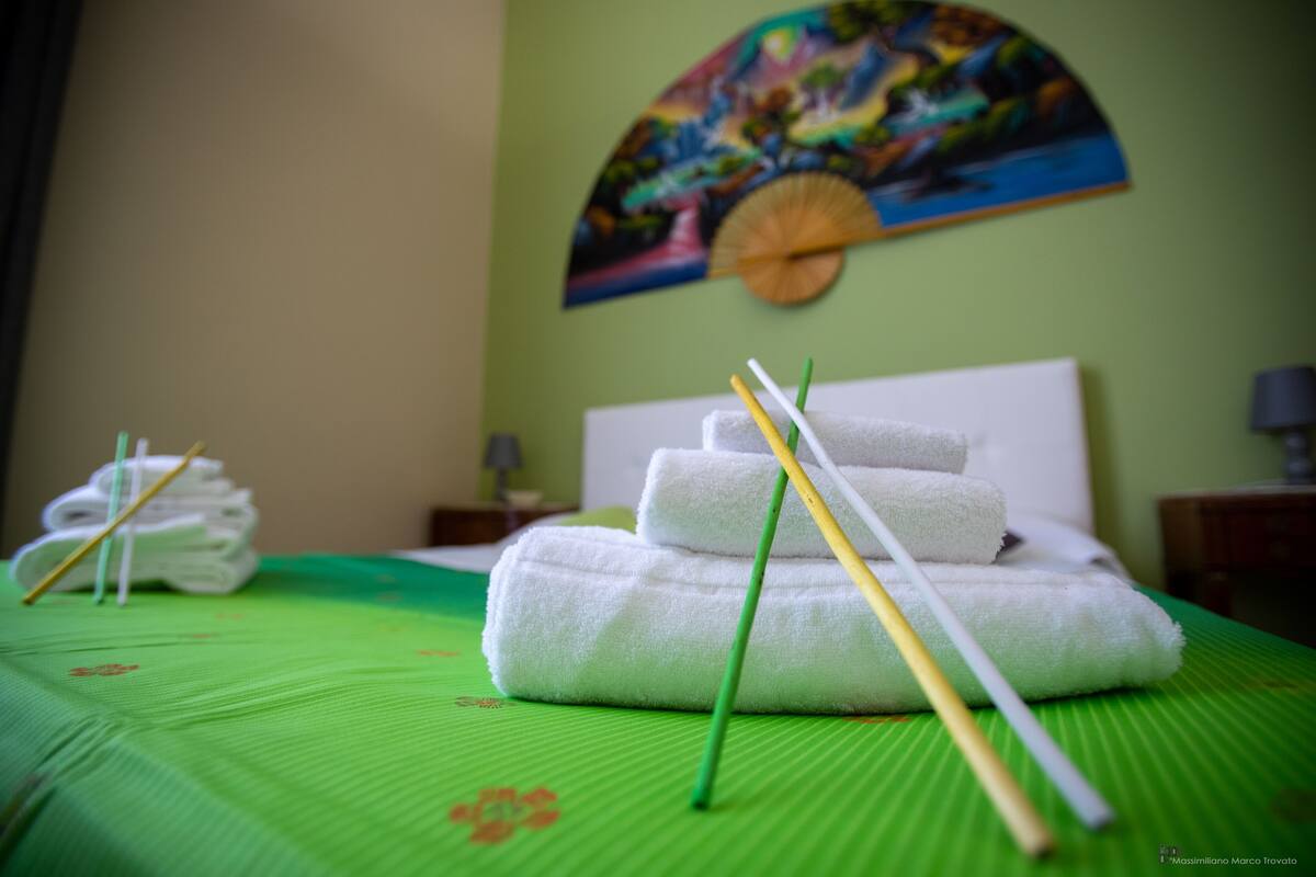 A neatly made bed with white towels and a green bedspread, a painting on the wall, and a small table with a lamp.
