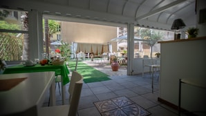 Un área de comedor al aire libre techada con mesas y sillas, un césped verde y vista del exterior del edificio.