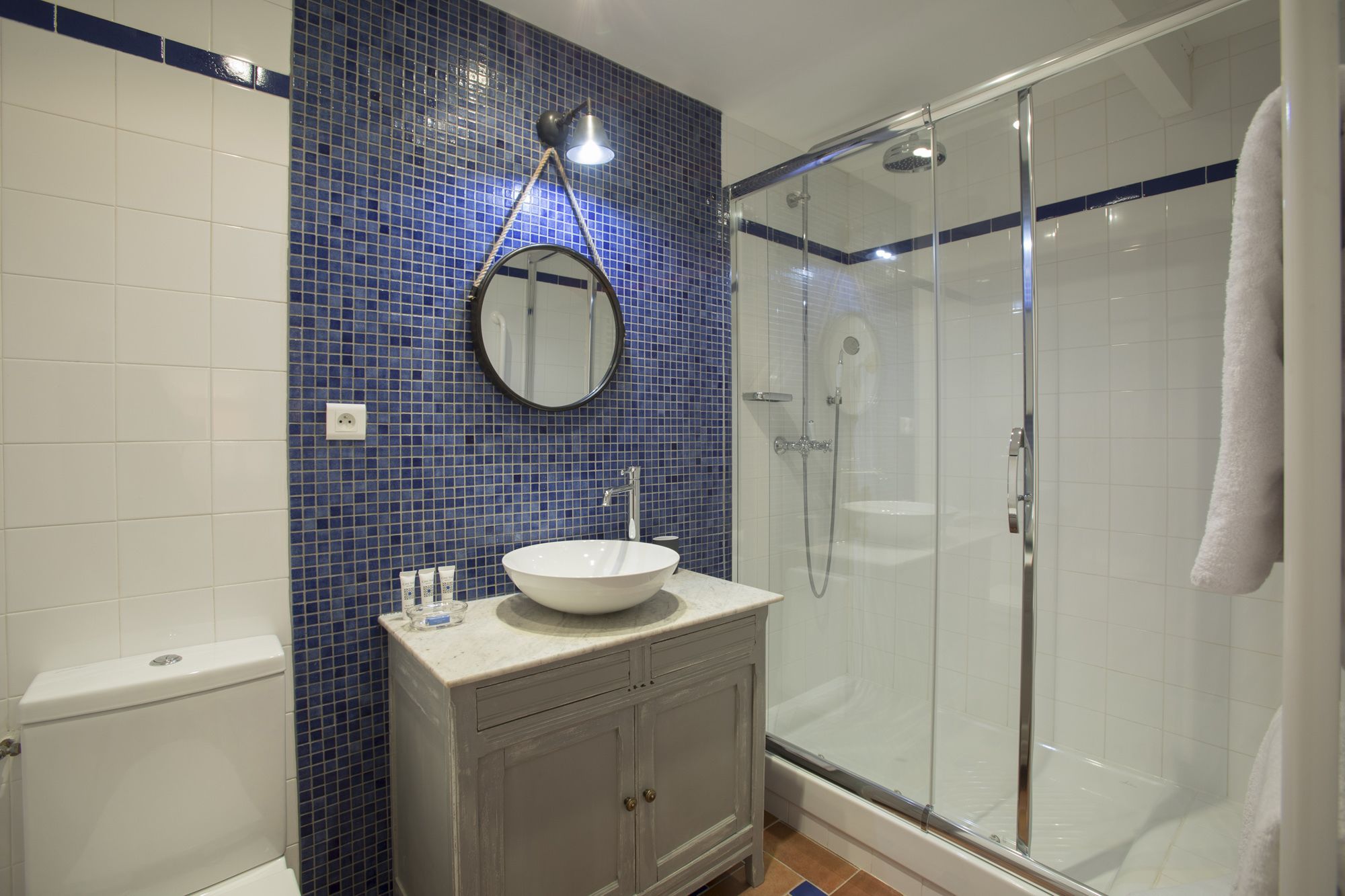 Un baño con inodoro blanco, lavabo con espejo redondo y ducha con cerramiento de vidrio.