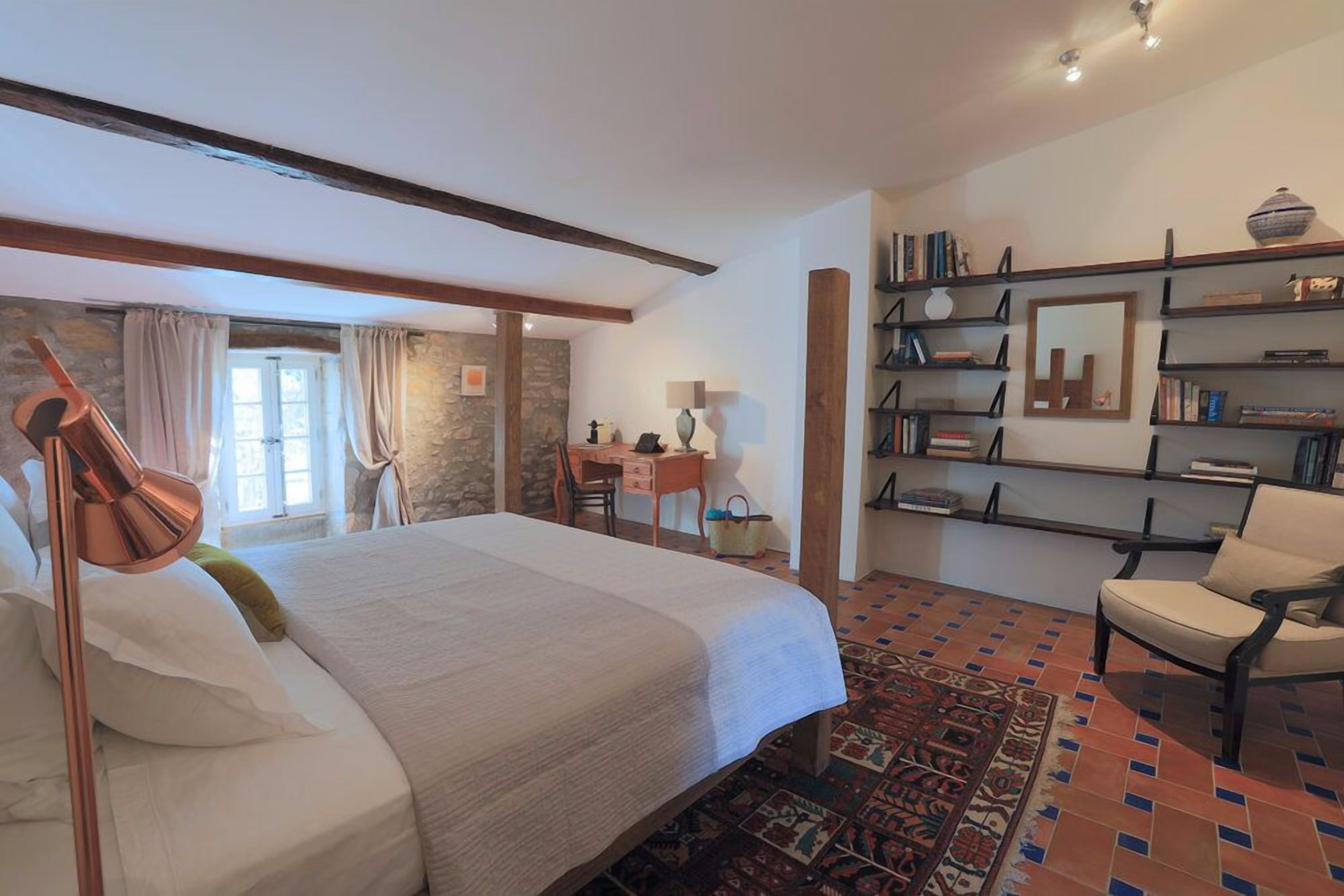 A bedroom with a large bed, a desk, a chair, a bookshelf, and a patterned rug.