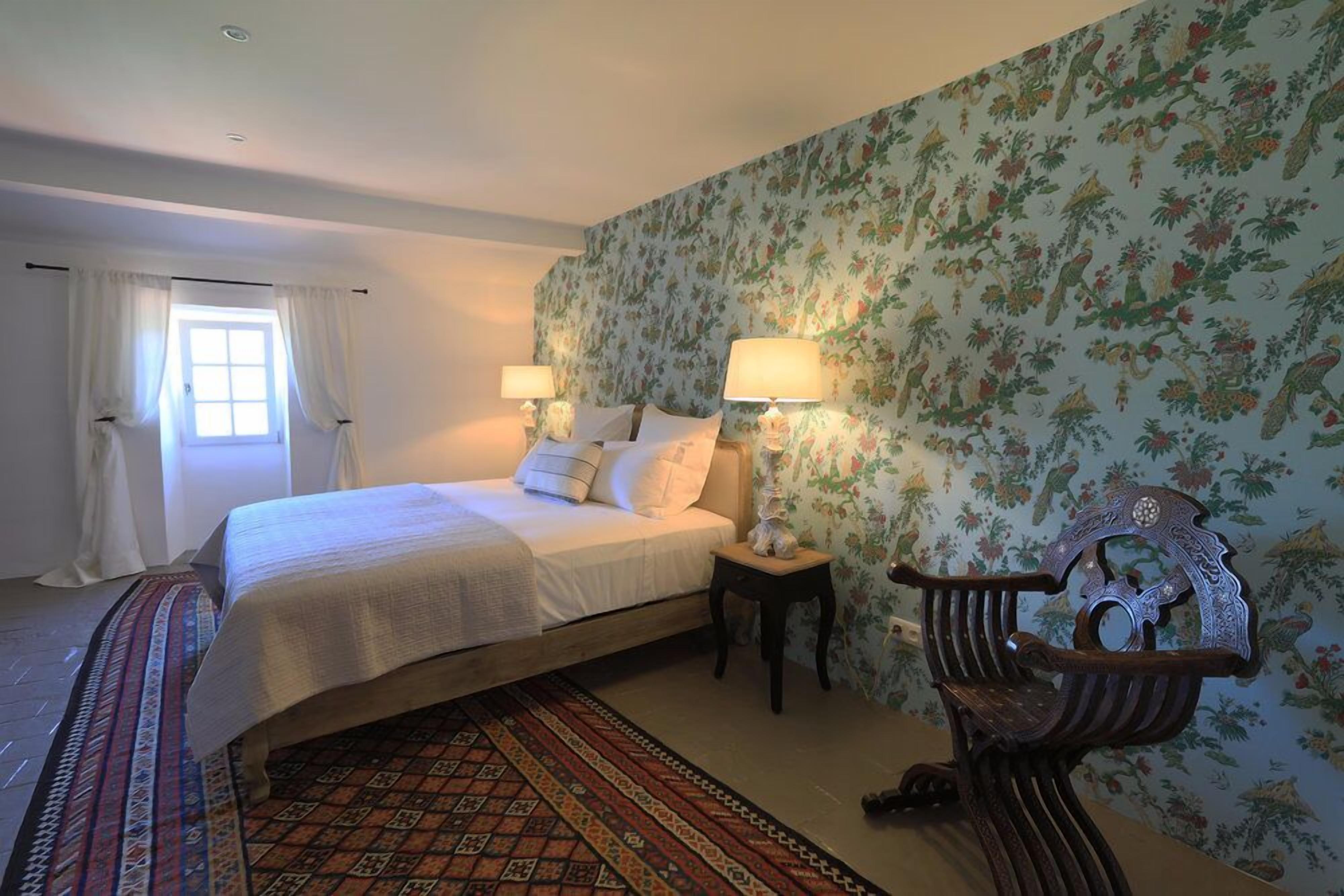 A bedroom with a bed, two bedside lamps, a chair, a window with curtains, and a floral wallpaper.