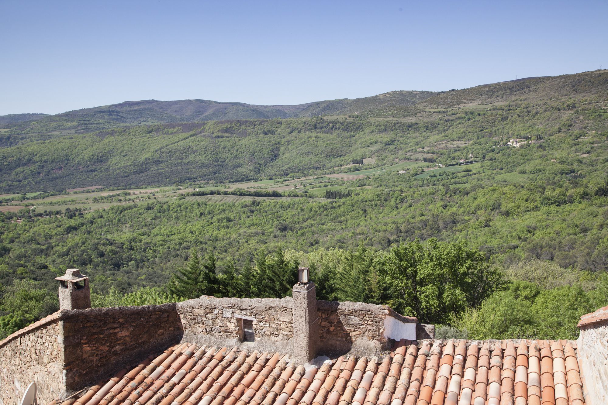A rustic stone building with terracotta roof tiles, overlooking a lush valley with scattered houses and dense greenery.
