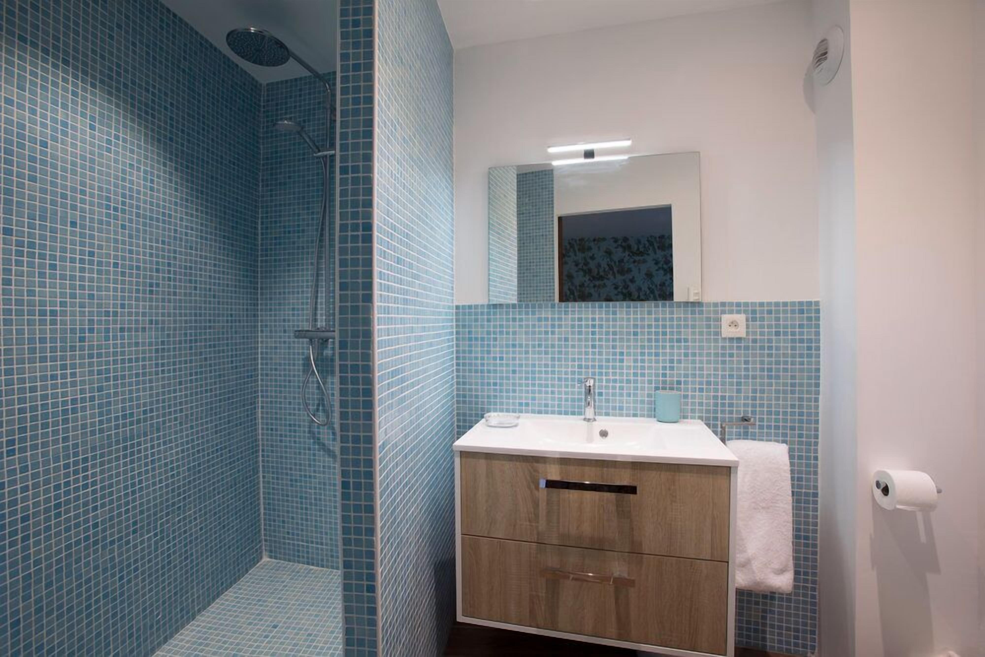 Une salle de bain avec des murs recouverts de carreaux bleus, un meuble-lavabo en bois, un lavabo blanc, un miroir et une douche.