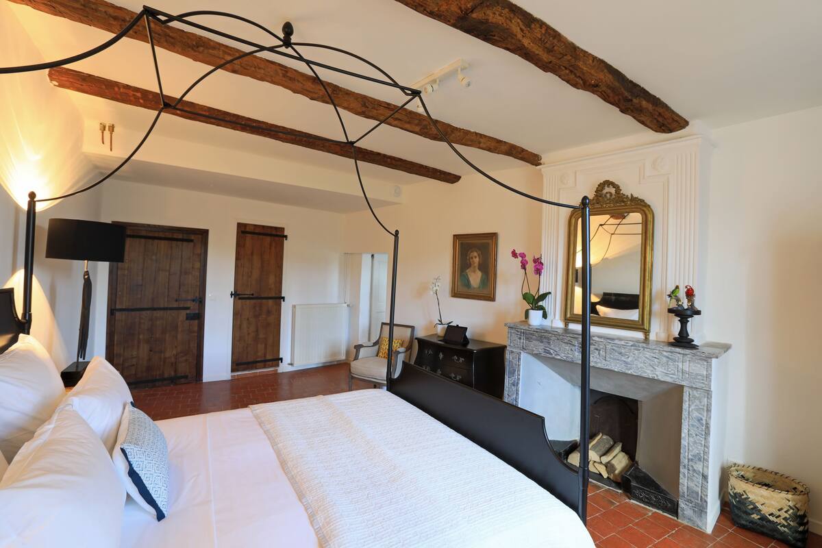 A spacious bedroom with a large bed, a fireplace, and a wooden ceiling beam.