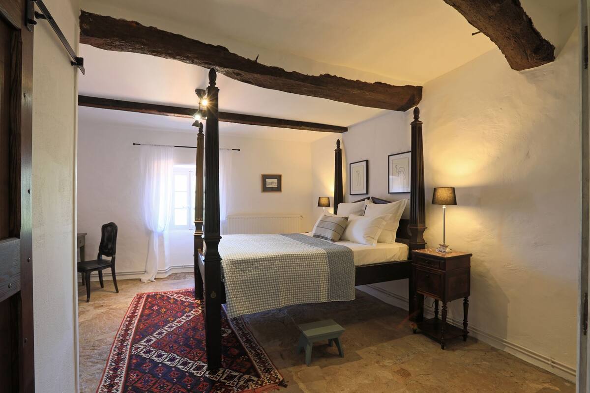 A bedroom with a four-poster bed, a small stool, a nightstand, and a patterned rug.