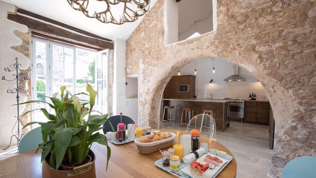 A breakfast setup with a stone archway leading to a kitchen, a wooden dining table with breakfast items, and a potted plant. - Chateau d'Olmet Guesthouse (Olmet-et-Villecun)