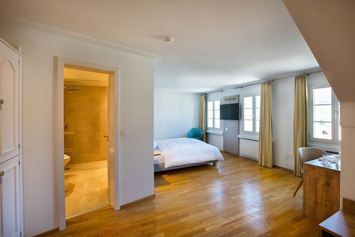 A spacious bedroom with a wooden floor, a bed, a desk, and a window with curtains.