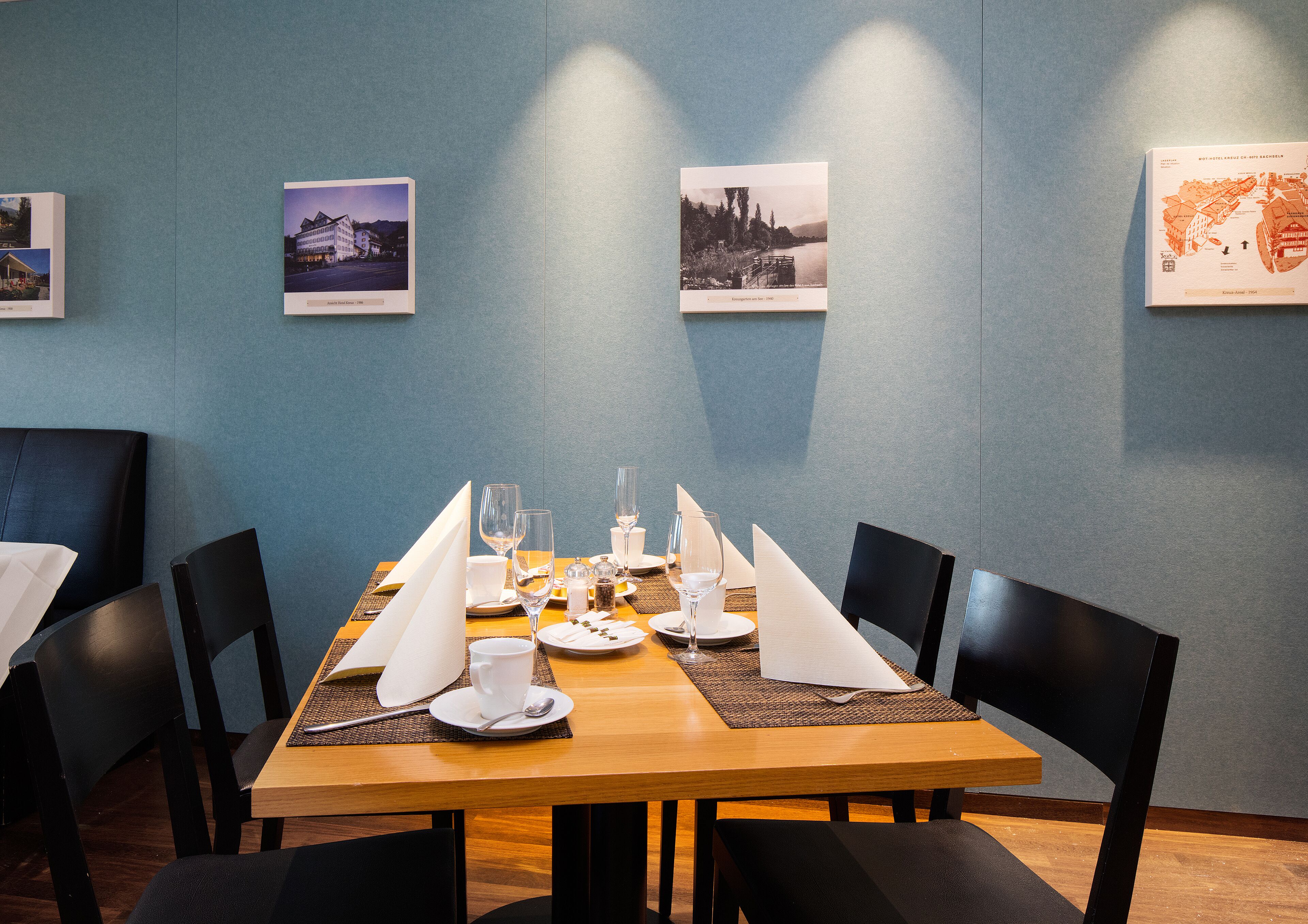 Un comedor con una mesa de madera dispuesta para cuatro personas, rodeada de sillas negras. Hay cuadros en la pared, un mapa y un menú sobre la mesa.