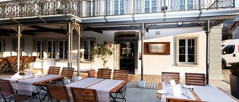 An outdoor café with wooden tables and benches, a sign reading "Kreuz," and a building with a balcony.