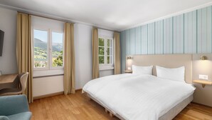 A bedroom with a bed, two windows with curtains, a desk, and a nightstand with a lamp. - Hotel Kreuz by b_smart (Sachseln)