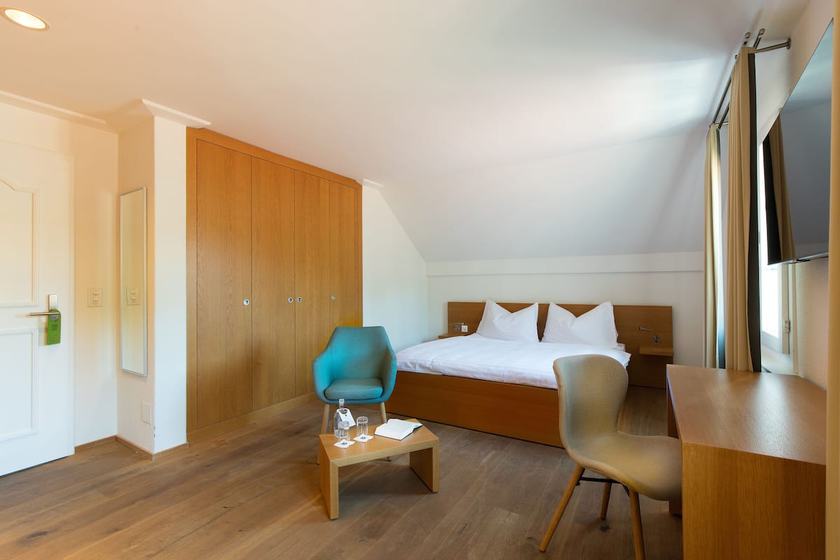 A bedroom with a bed, two chairs, a small table, wooden wardrobe, and a TV.
