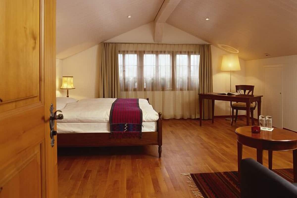 A hotel room with a wooden floor, a bed with a red blanket, a desk with a chair, and a window with curtains.