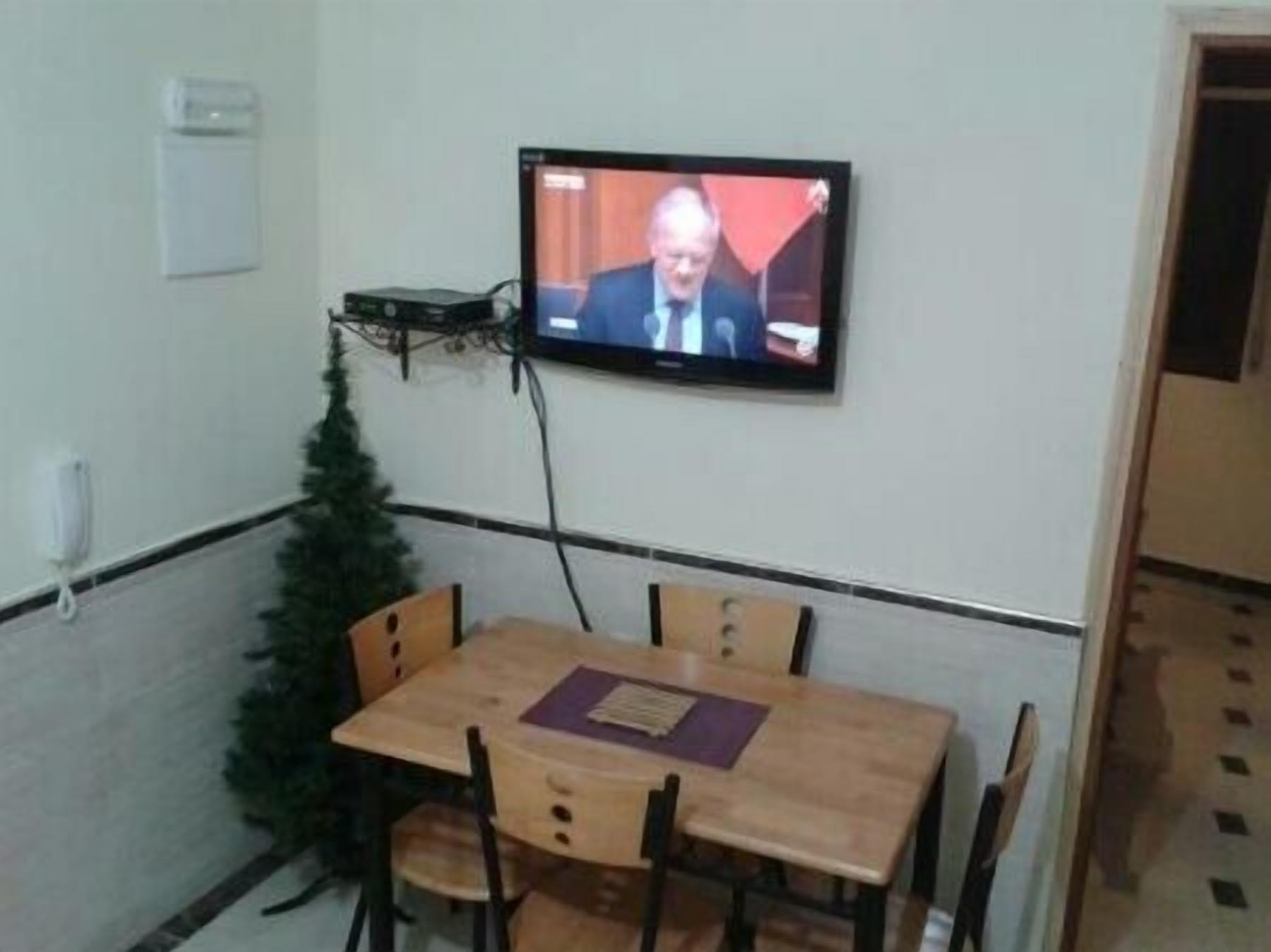 Een kamer met een aan de muur bevestigde televisie, een kleine kerstboom en een houten tafel met stoelen.