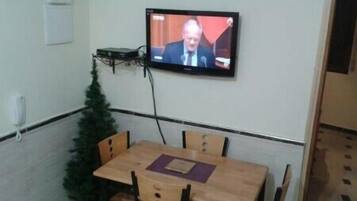 Een kamer met een aan de muur bevestigde televisie, een kleine kerstboom en een houten tafel met stoelen.