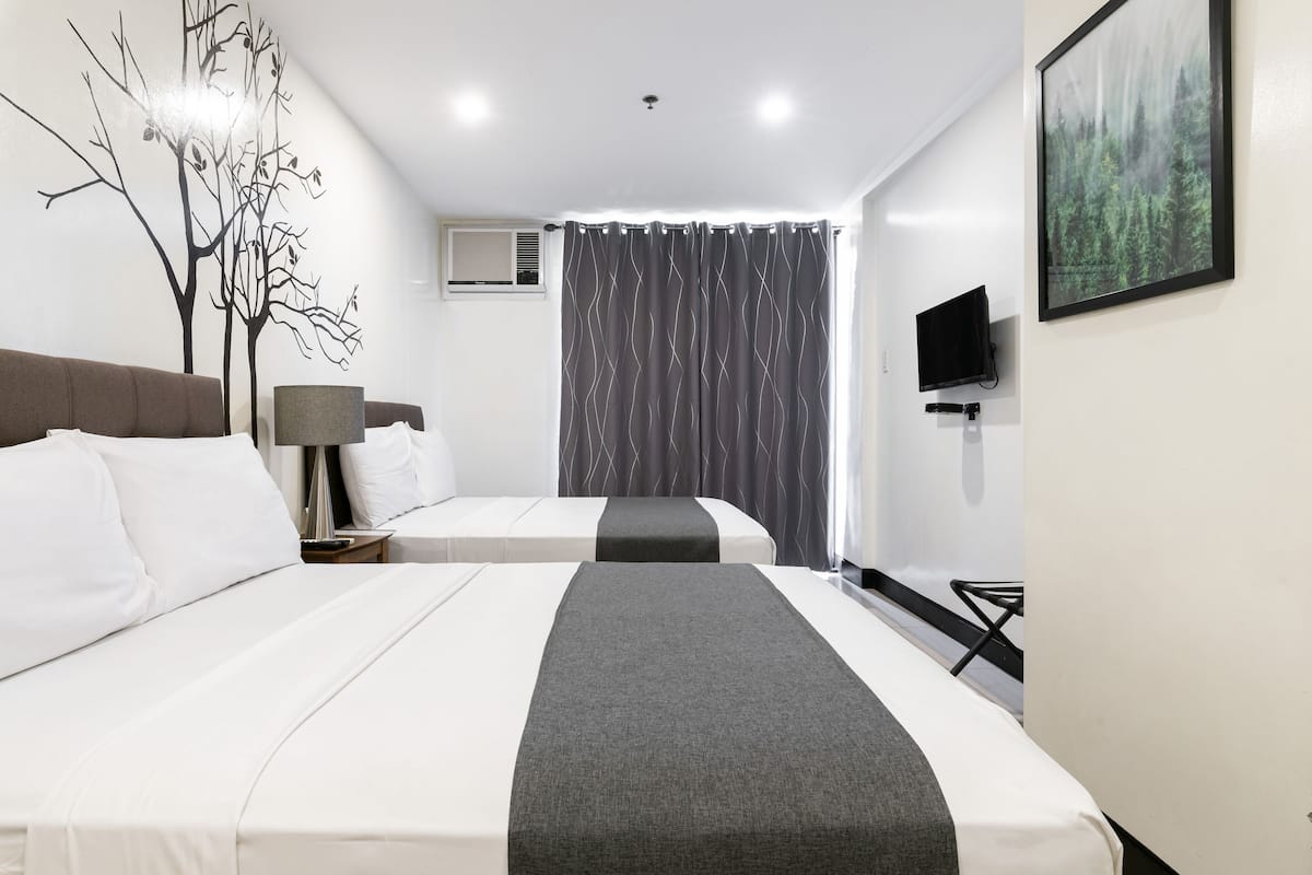 A hotel room with two beds, a flat-screen TV, a wall-mounted air conditioner, and a large window with curtains.