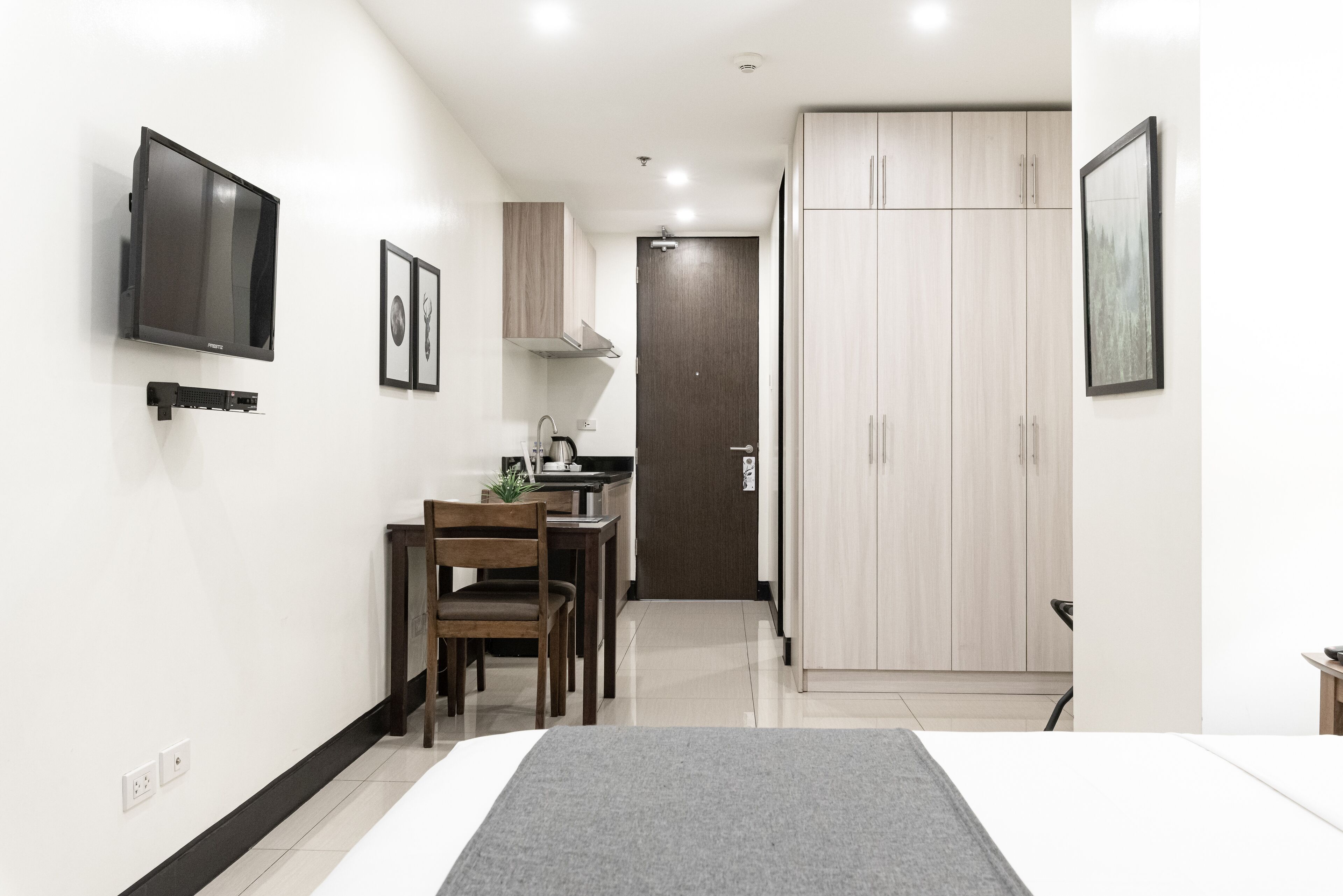 A compact hotel room with a bed, a desk with a chair, a TV mounted on the wall, and a wardrobe.