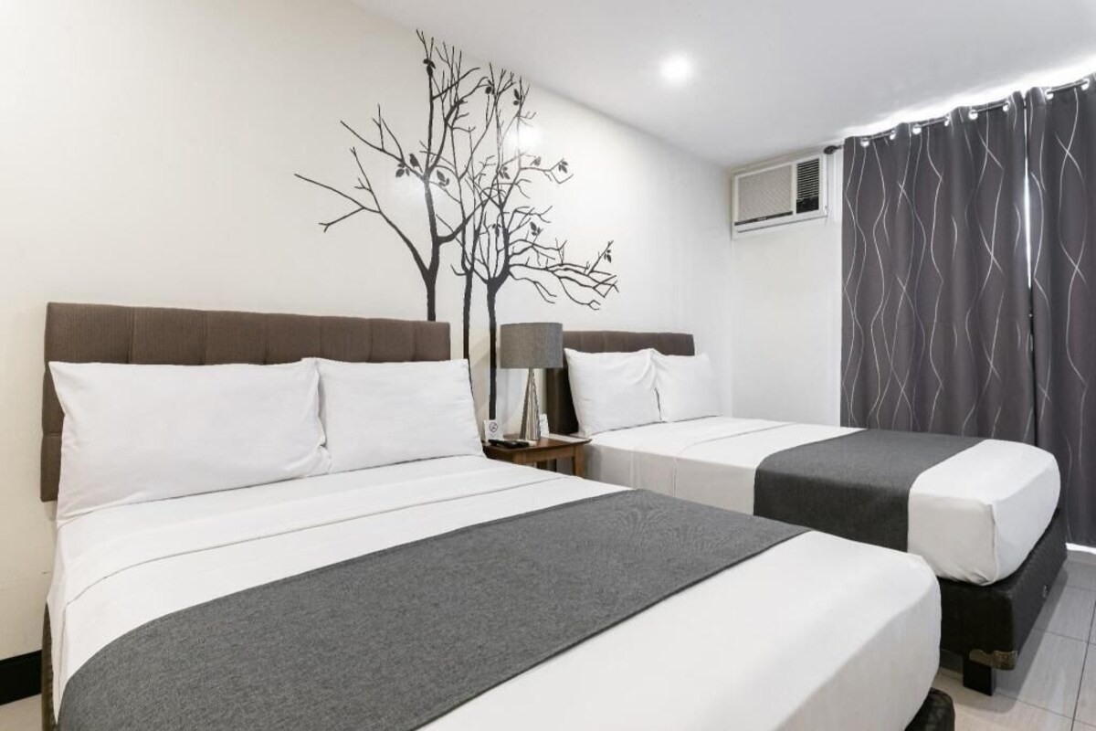 Habitación de hotel con dos camas, una mesita de noche, una lámpara, un mural con motivos de árboles y una cortina.
