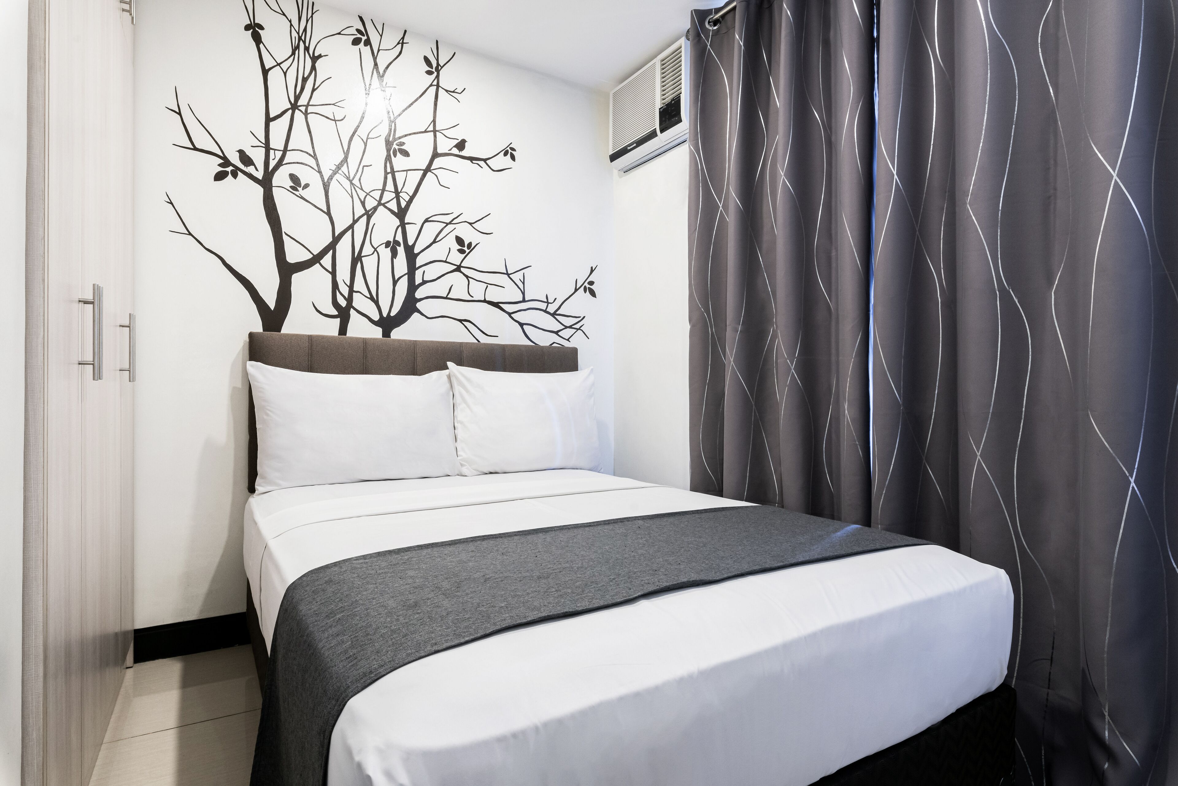 A neatly made bed with white and gray bedding, a tree mural on the wall, and a gray curtain with a wavy pattern.