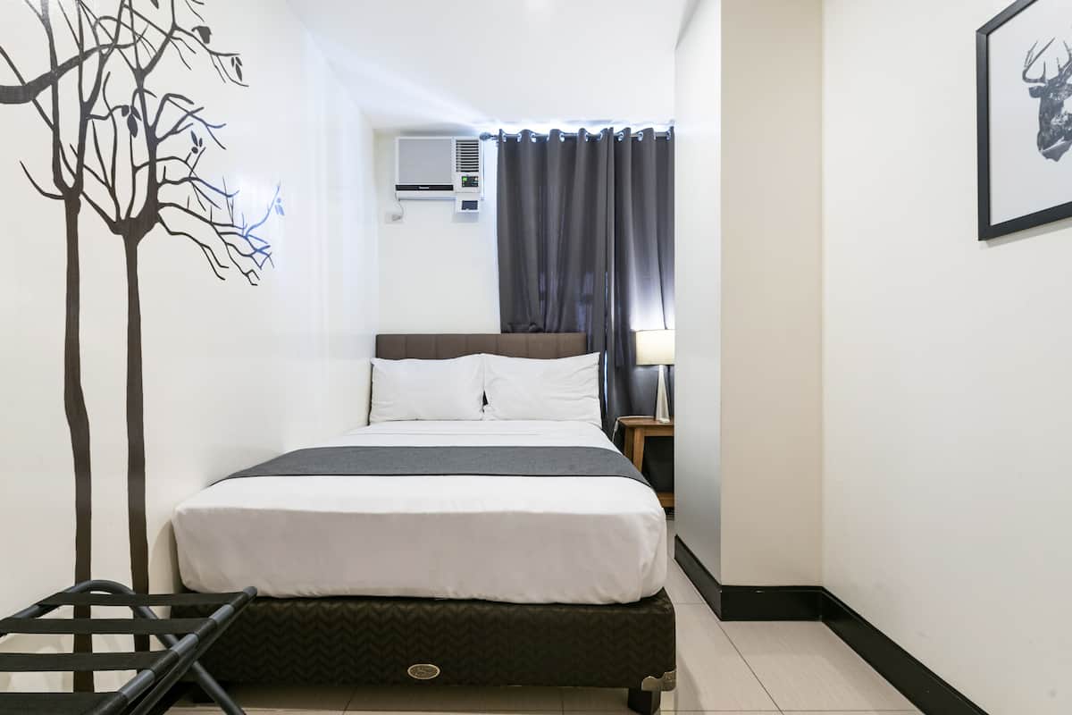 A hotel room with a bed, a nightstand, a lamp, an air conditioning unit, and a wall-mounted artwork.