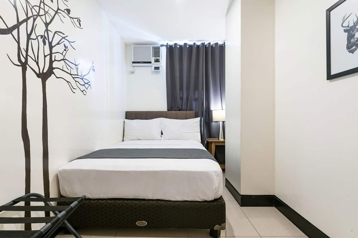 A hotel room with a bed, a nightstand, a lamp, an air conditioning unit, and a wall with a tree design.