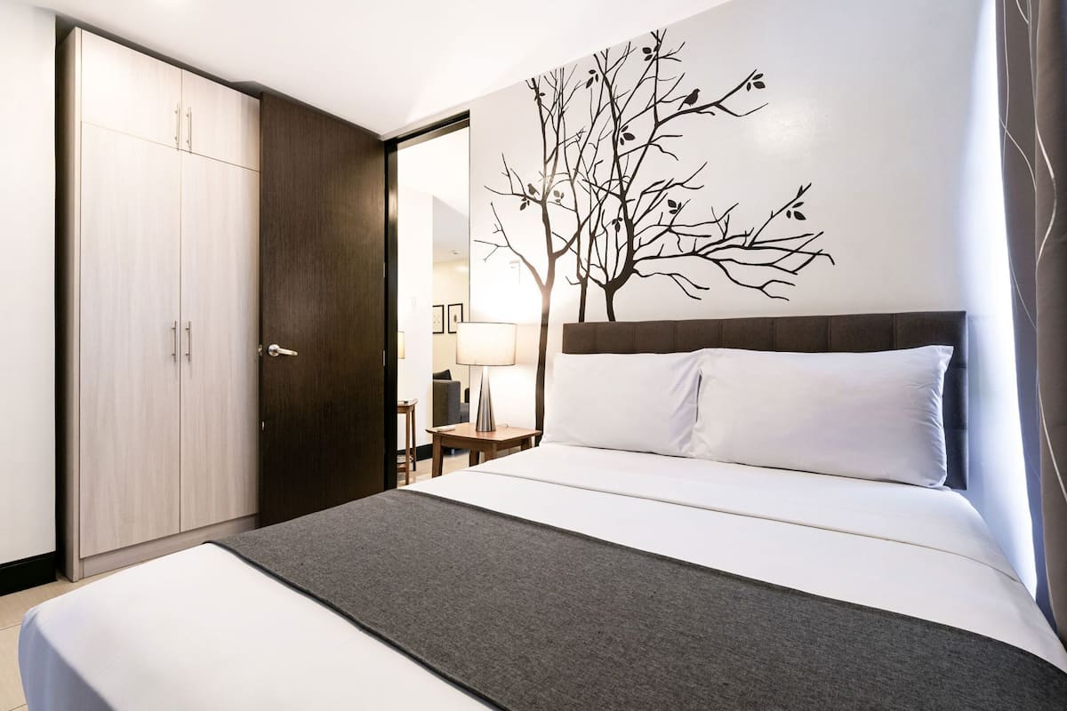 A hotel room with a bed, a wardrobe, a nightstand, a lamp, and a wall mural.